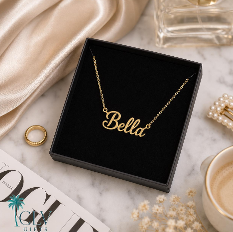 Gold-tone Bella script name necklace in a black jewelry box on a cream styled surface.